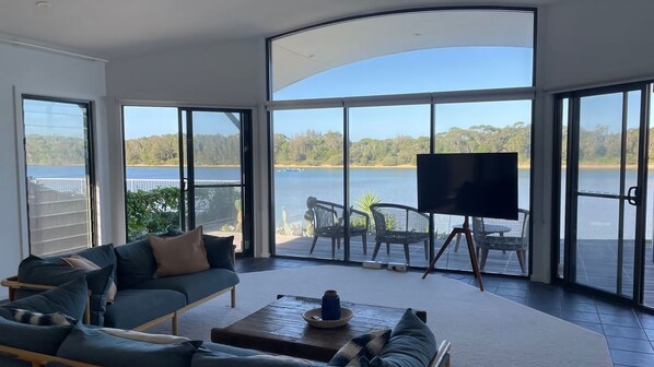 A spacious living area with a large window overlooking a lake, furnished with a sofa, chairs, and a coffee table. - Absolute Waterfront Beach House (Minnamurra)