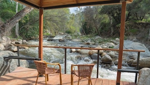 A wooden deck with wicker chairs overlooking a flowing river with rocks. - The River's Edge - Private Home with River Access, Spa, and Karaoke (Three Rivers)