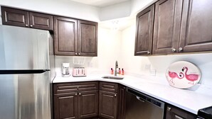 Cocina compacta con armarios de madera oscura, refrigerador de acero inoxidable, lavavajillas y encimera blanca.