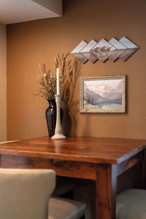 A dining table with a vase of dried plants and a candle, a framed landscape painting, and a geometric wall decoration.