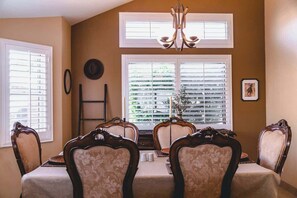 Une salle à manger avec une table dressée pour quatre personnes, flanquée de deux fenêtres équipées de stores, d’un lustre et d’une échelle appuyée contre le mur.