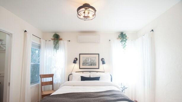A bedroom with a bed, a chair, a hanging plant, a framed picture, and a ceiling light fixture.