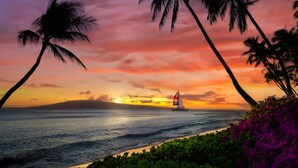 A sunset over the ocean with a sailboat, palm trees, and a beach.