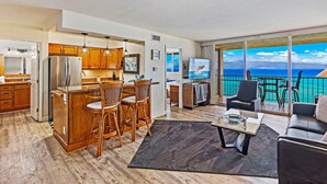A modern kitchen with wooden cabinets and a bar area, a dining set, and a living area with a view of the ocean.