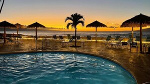 A pool area with lounge chairs and umbrellas, overlooking the ocean at sunset.