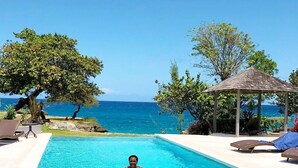 Uma piscina azul cristalina com uma pessoa nadando, rodeada por cadeiras de descanso e um gazebo, com vista para o oceano.
