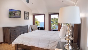 Um quarto com uma cama, uma mesa de cabeceira, uma luminária, uma cadeira, uma janela com vista para o mar e um quadro na parede.