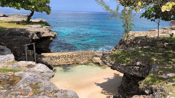 Paisagem costeira com praia clara, formações rochosas e vista para o oceano.