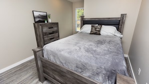 A bedroom with a wooden bed frame, a marble-patterned bedspread, a nightstand with drawers, a framed picture on the wall, and a window with a view of trees. - Quiet and cozy new townhome minutes from the mall exit. (Barboursville)