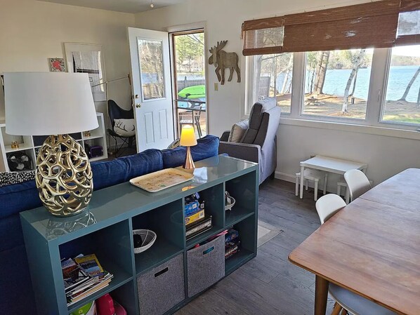 Una sala de estar acogedora con un sofá azul, una mesa de centro de madera y una lámpara. Hay una estantería llena de libros y una pequeña mesa con una silla. Una puerta da a una terraza con vista al agua.