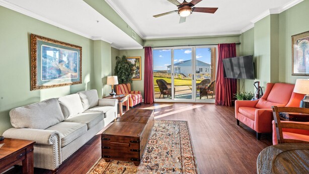 A spacious living room with a large sofa, a coffee table, and a television. There is a ceiling fan and a view of a house through the glass doors. - 3BR Fort Morgan Condo | Pool & Hot Tub (GULF SHORES)