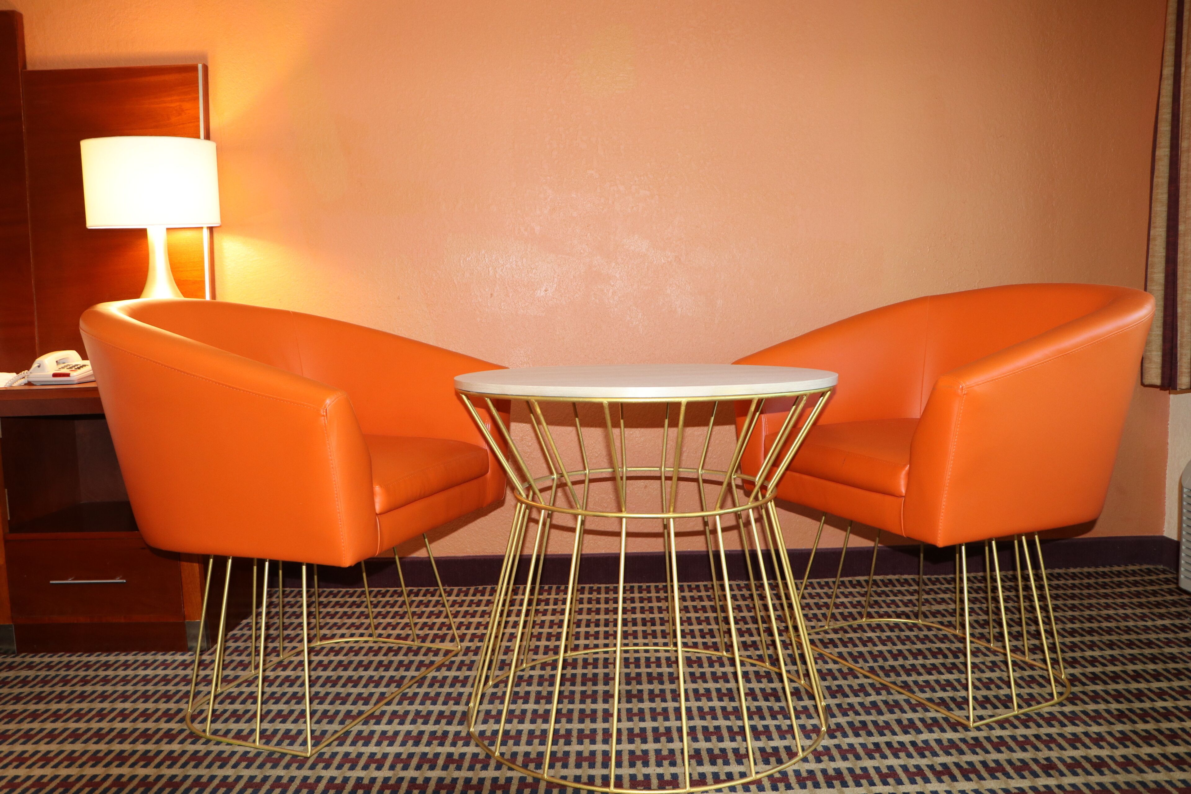 Habitación de hotel con dos sillones naranjas, una mesa redonda pequeña, una lámpara y una alfombra estampada.
