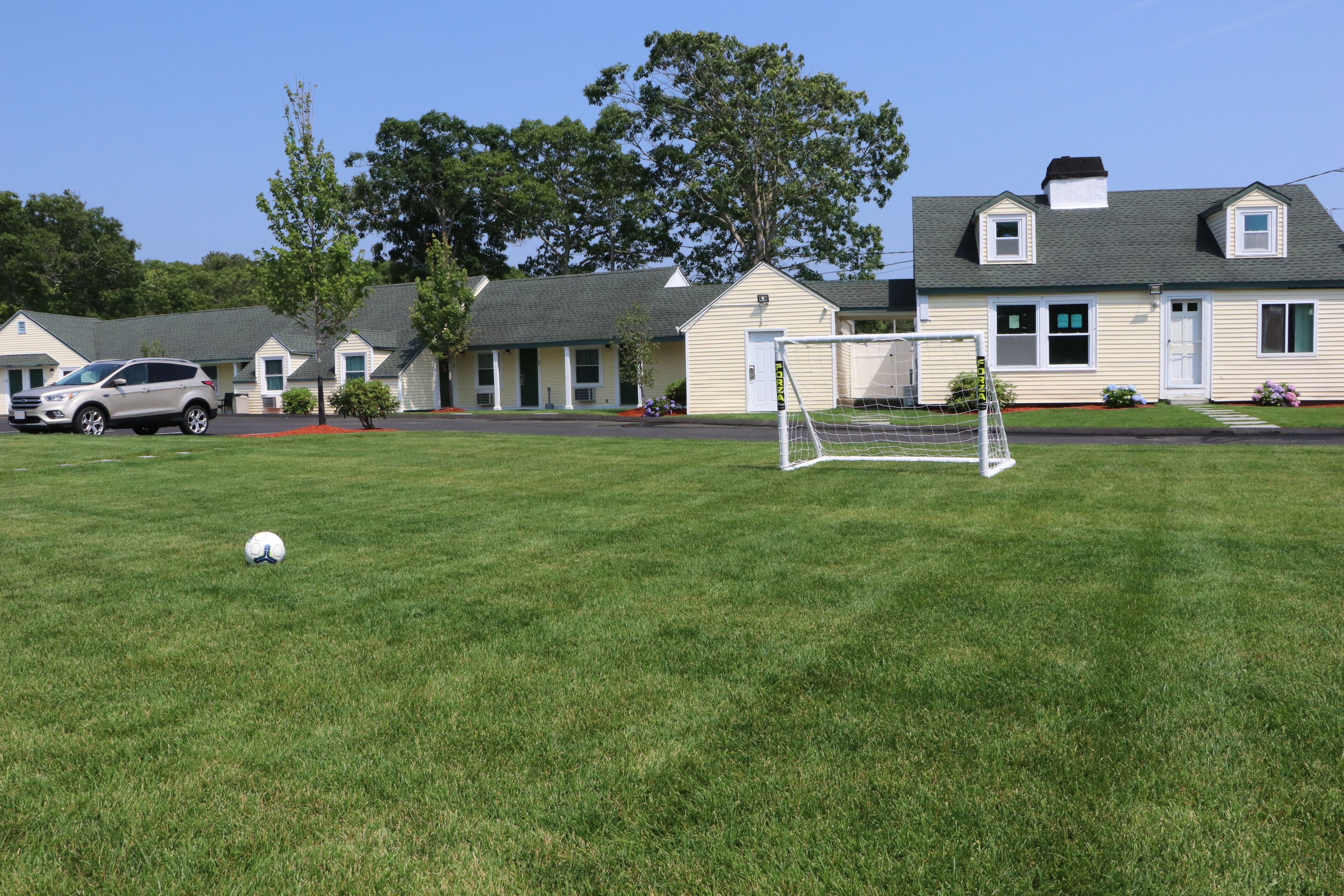 Um campo gramado com um gol de futebol, um carro e uma fileira de edifícios no estilo motel ao fundo.