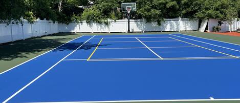 Una cancha de tenis azul con un aro de baloncesto, rodeada por una valla blanca y árboles.