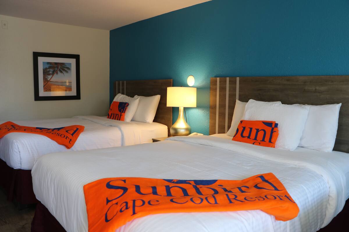 A hotel room with two beds, each with a white bedspread and orange towels. There is a framed picture on the wall and a lamp on the nightstand.