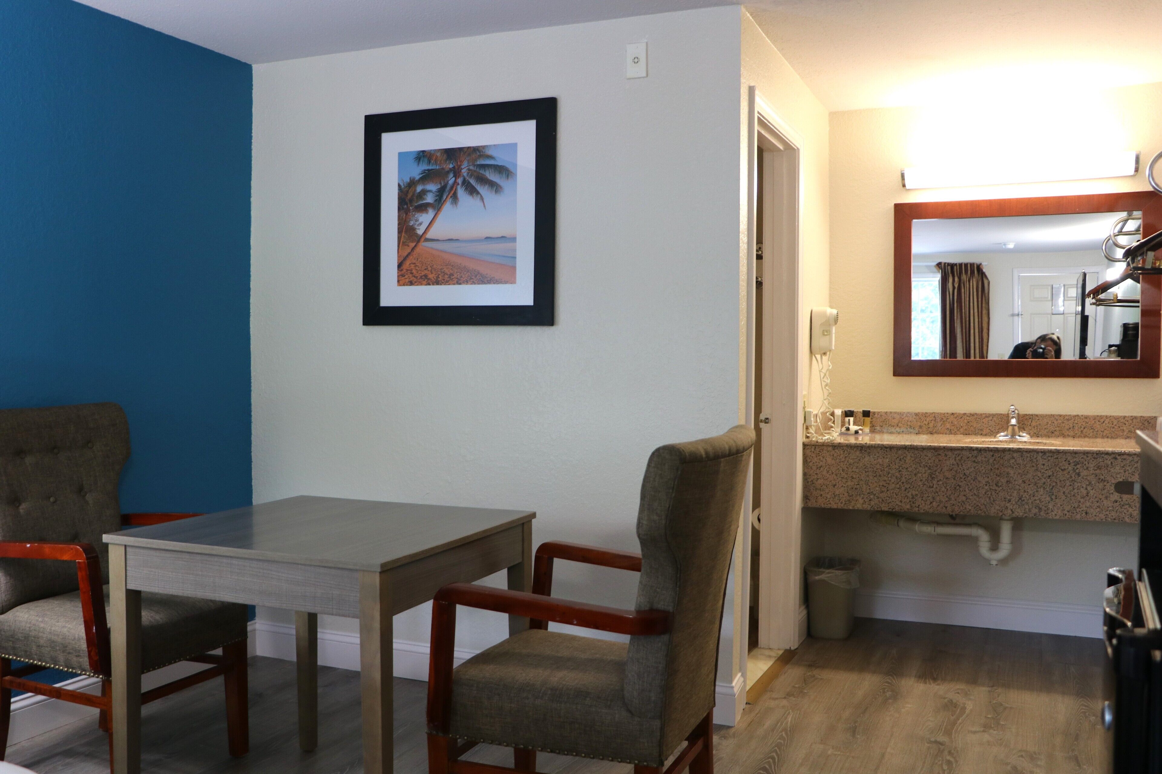A hotel room with a dining area, a bathroom with a sink, and a framed picture of a beach scene.