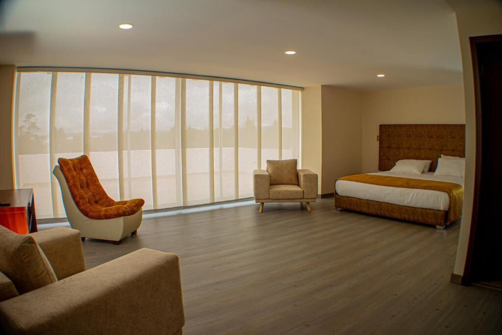 A modern hotel room with a large window, a bed, two armchairs, and a small table.