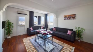 Ein modernes Wohnzimmer mit einer Couch, einem Esstisch für zwei Personen und Blick auf die Stadt durch große Fenster.