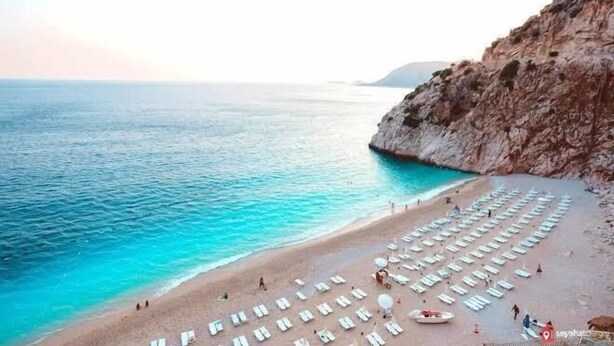 Ein Strand mit Reihen von Liegestühlen und Sonnenschirmen, eine felsige Klippe und ruhiges türkisfarbenes Wasser.
