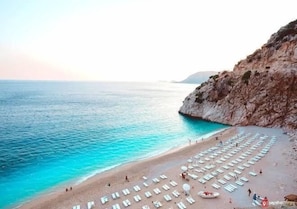 A beach with rows of sun loungers and umbrellas, a rocky cliff, and calm turquoise waters. - Bilginer Apart (Demre)