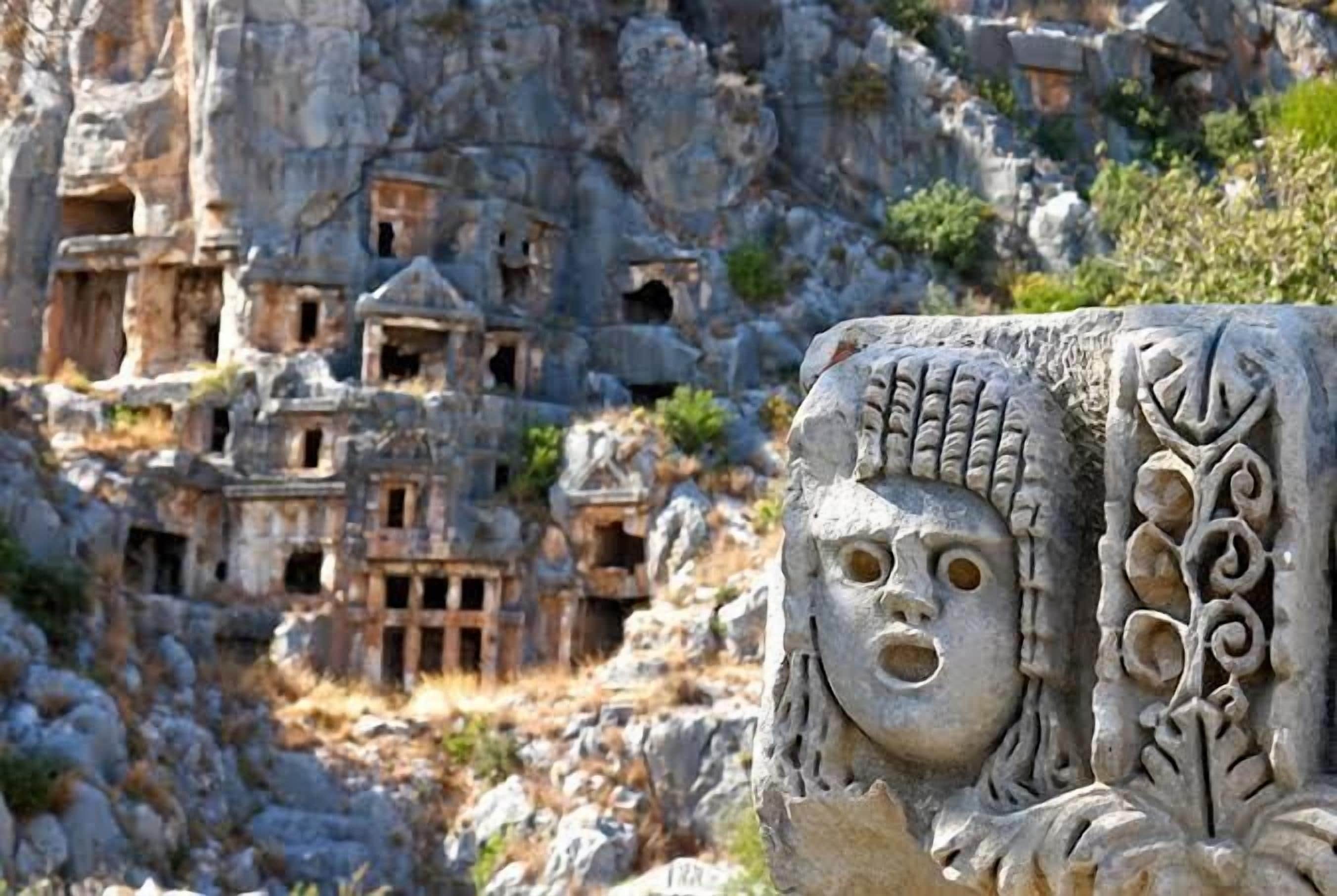 A carved stone face with intricate designs, set against a backdrop of ancient rock-cut buildings.