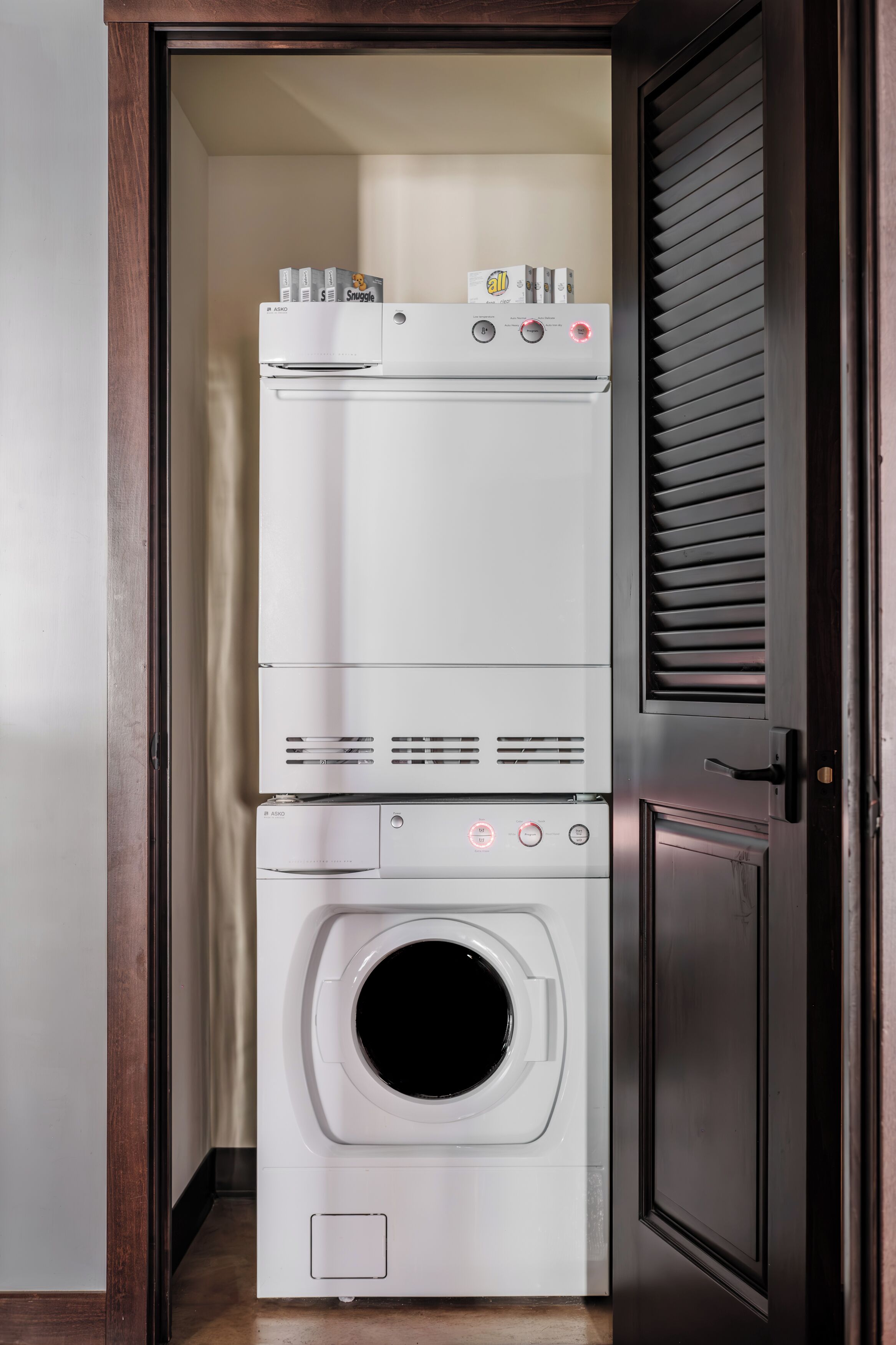 A compact laundry room with a stacked washer and dryer, a door with a handle, and a wooden frame around the door.