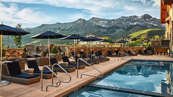 A poolside area with lounge chairs and umbrellas, a stone wall, and a view of mountains.