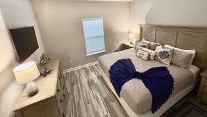 Un dormitorio con cama, mesitas de noche, ventilador de techo, ventana con persianas y televisor en la pared.