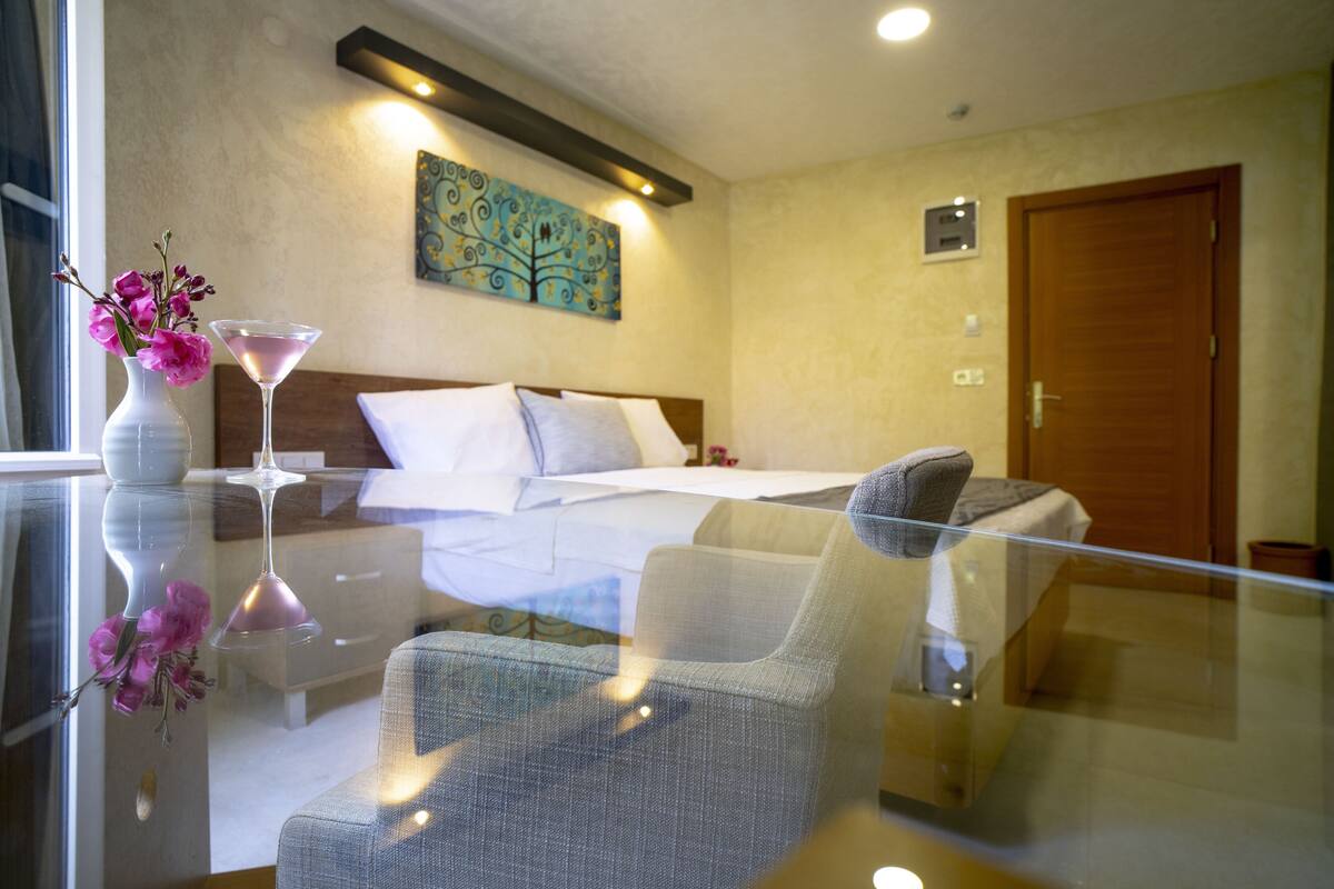 A modern hotel room with a glass table, a bed with white linens, a painting on the wall, and a wooden door.