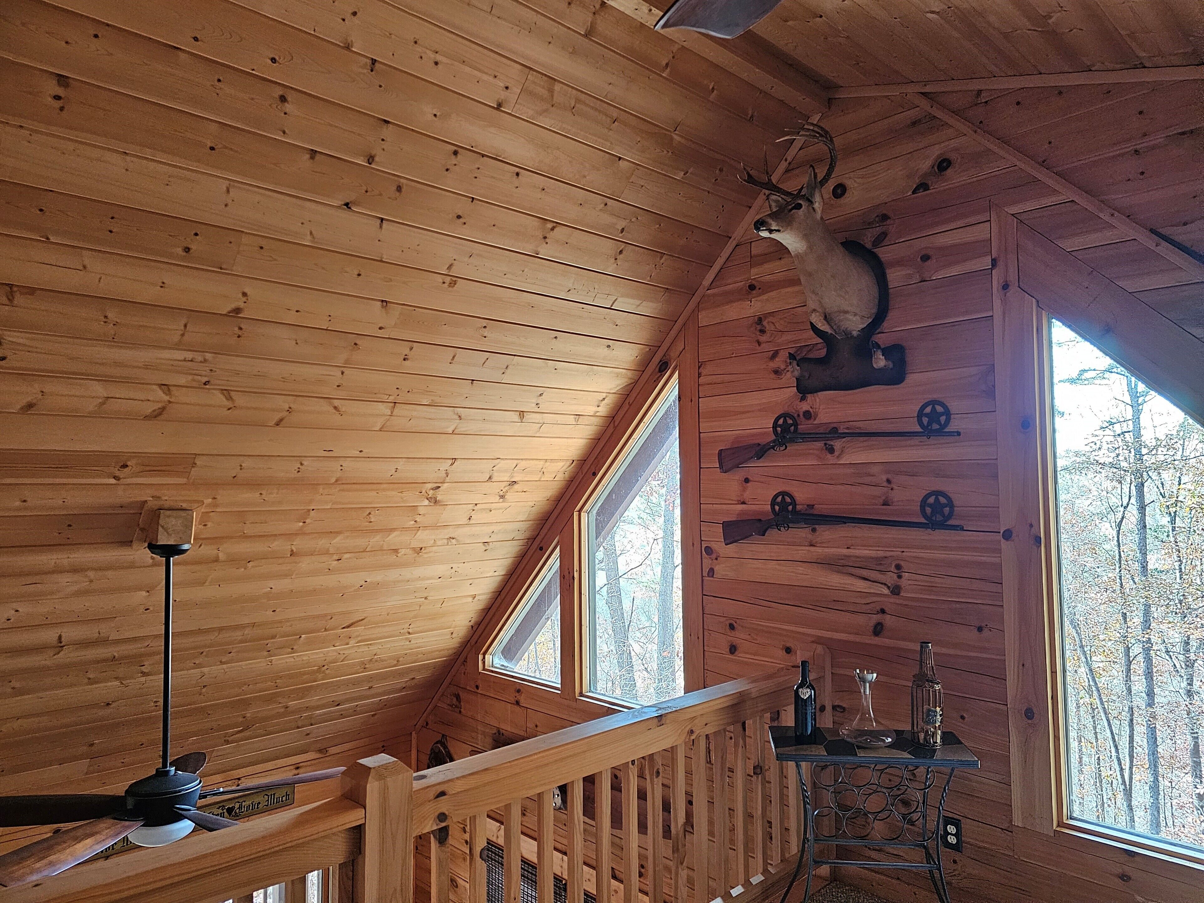 Interior de una cabaña de madera con ventilador de techo, cabeza de venado montada y una mesa con botellas.