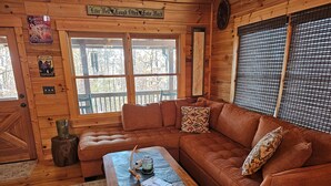 Ein gemütliches Blockhaus-Interieur mit Holzboden, braunem Ledersofa, Couchtisch und Fenster mit Blick auf Bäume.
