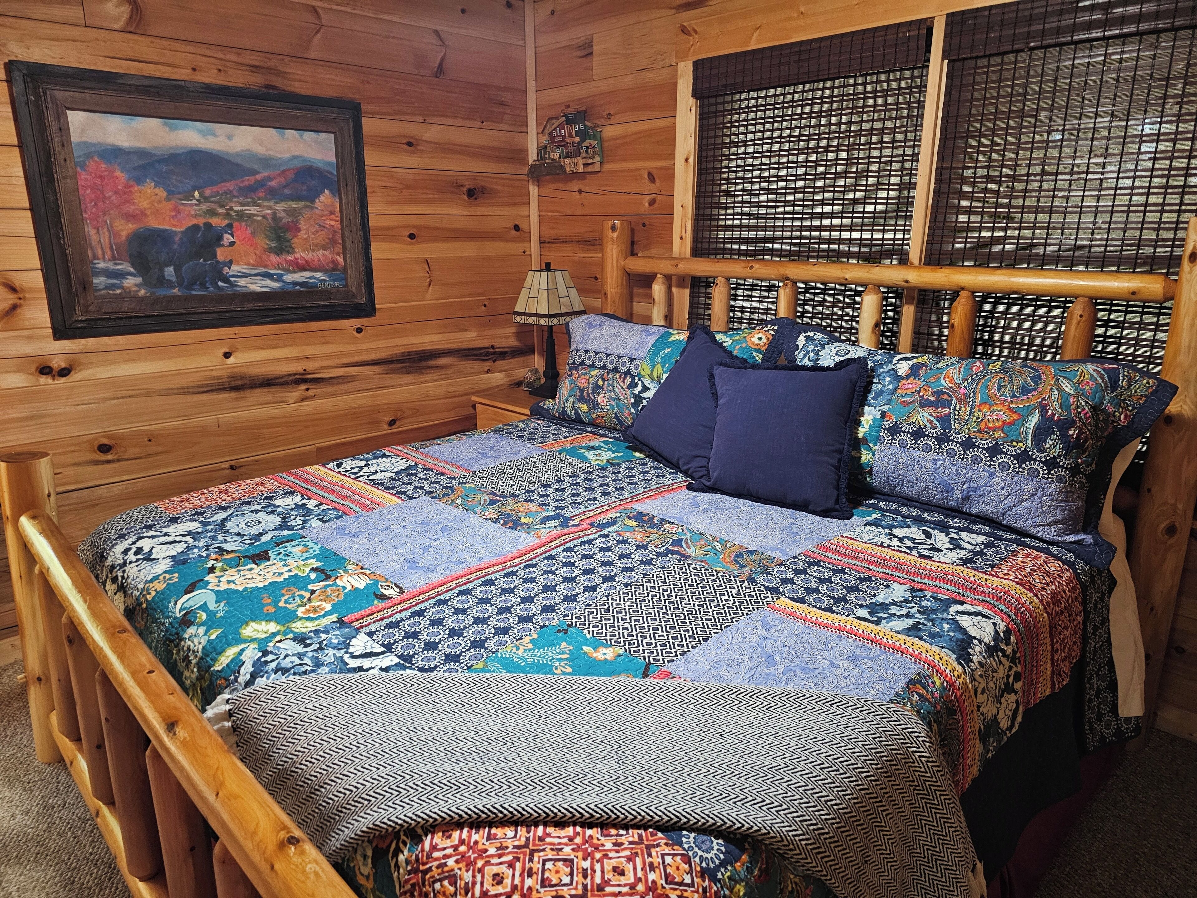 Ein Schlafzimmer einer Holzhütte mit einem Bett, das eine farbenfrohe Patchworkdecke zeigt, einem Bärenbild an der Wand und einem Fenster mit einem Insektenschutznetz.