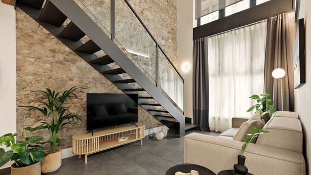 A modern living room with a stone wall, a staircase, a flat-screen TV, a sofa, and a coffee table. - OLA LIVING MACBA 0 (Barcelona)