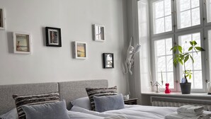 A neatly made bed with white linens and a grey headboard, a window with a view of greenery, and several framed pictures on the wall. - Spacious luxury apartment in city Center . (Copenhagen)