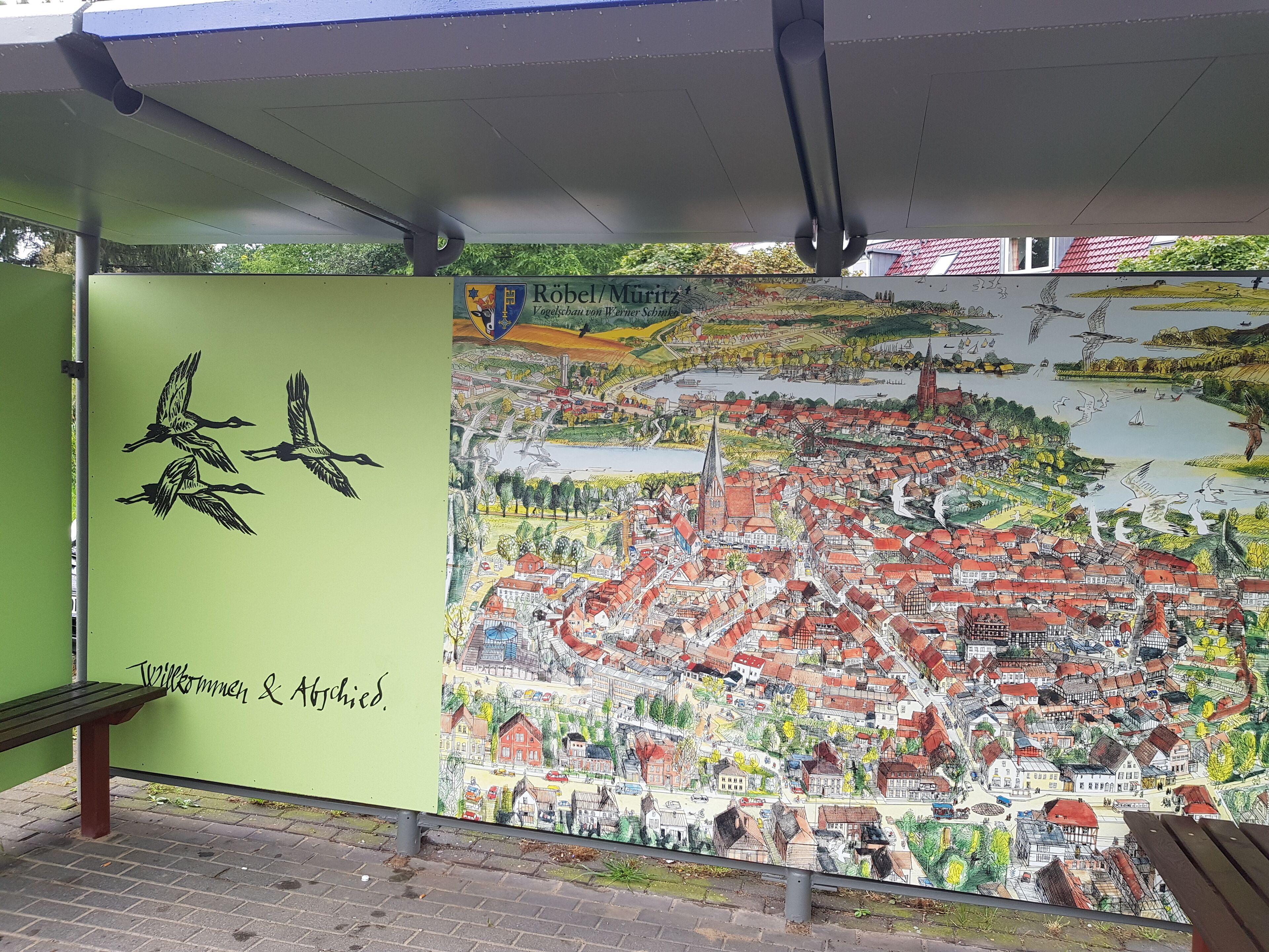 Ein detailliertes Wandgemälde zeigt eine Stadt mit einer Bank und einem Willkommensschild.