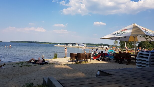 Une plage où des gens se prélassent au soleil et se baignent, un quai avec des bateaux, et un grand parasol.