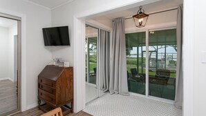 Une pièce avec un meuble en bois, un téléviseur fixé au mur et une grande porte-fenêtre coulissante donnant sur un espace extérieur avec vue sur l’eau et la verdure.