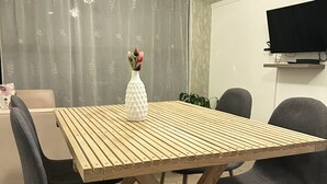 Un coin repas avec une table en bois, quatre chaises, un vase de fleurs, un canapé, un téléviseur et un rideau.