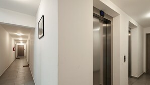 Un couloir moderne avec un ascenseur central, un extincteur et un tableau encadré accroché au mur.