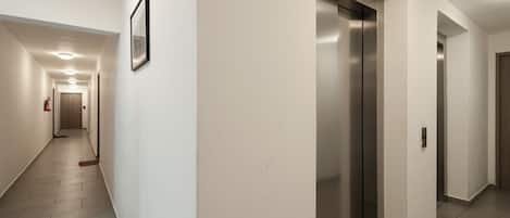 A modern hallway with a central elevator, a fire extinguisher, and a framed picture on the wall.
