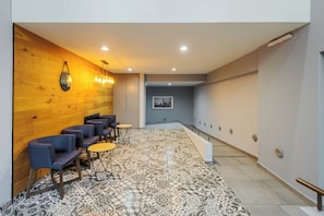 A hotel lobby with a long corridor, seating area with chairs and tables, wooden wall, and patterned floor tiles.