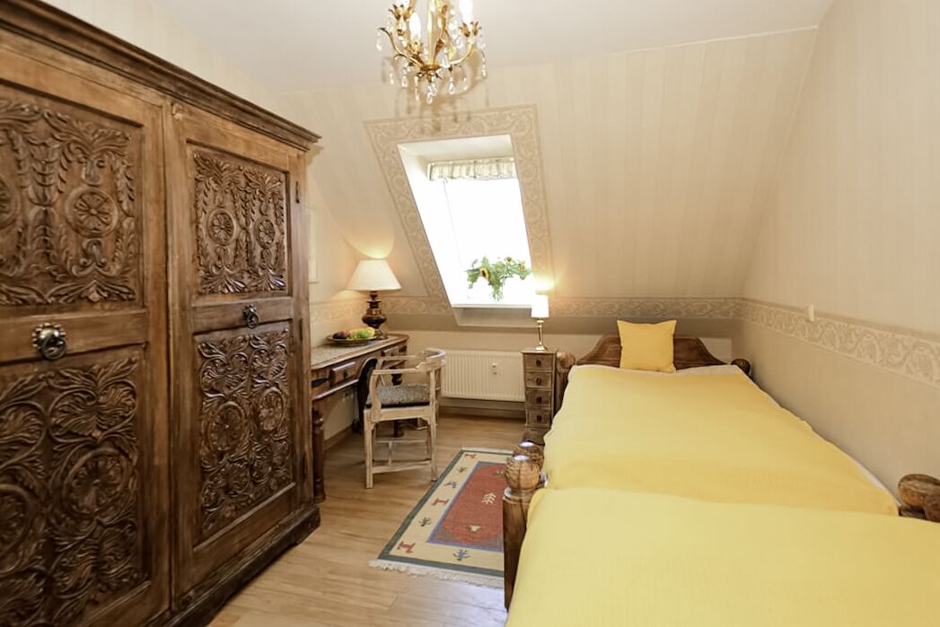 A bedroom with a wooden wardrobe, a desk, a chair, a bed with yellow bedding, a nightstand, a lamp, and a window with a plant.