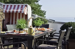 Un área de comedor al aire libre con mobiliario de mimbre, un toldo a rayas y plantas en macetas.