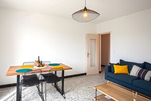 Uma sala de estar moderna com uma mesa de jantar de madeira, cadeiras pretas com estrutura de arame, um sofá azul com almofadas amarelas e listradas, e um lustre.
