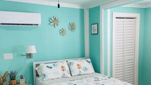 Een slaapkamer met een bed, een plafondventilator, een airconditioner, een nachtkastje, een lamp, een klein plankje met decoratieve spullen, en een pluizig, wit tapijt.
