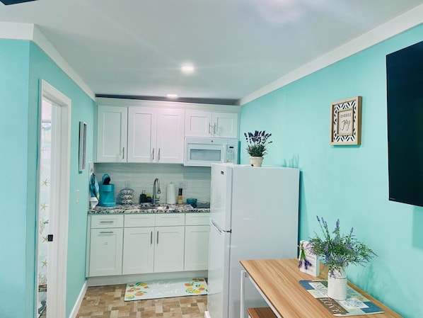 Una cocina compacta con armarios blancos, microondas, refrigerador y un pequeño comedor con mesa de madera.