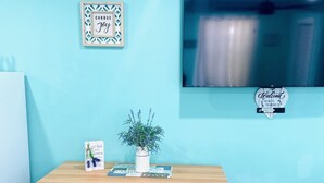 Een kitchenette met een houten tafel, twee krukken, een plant in pot en een aan de muur bevestigde televisie.
