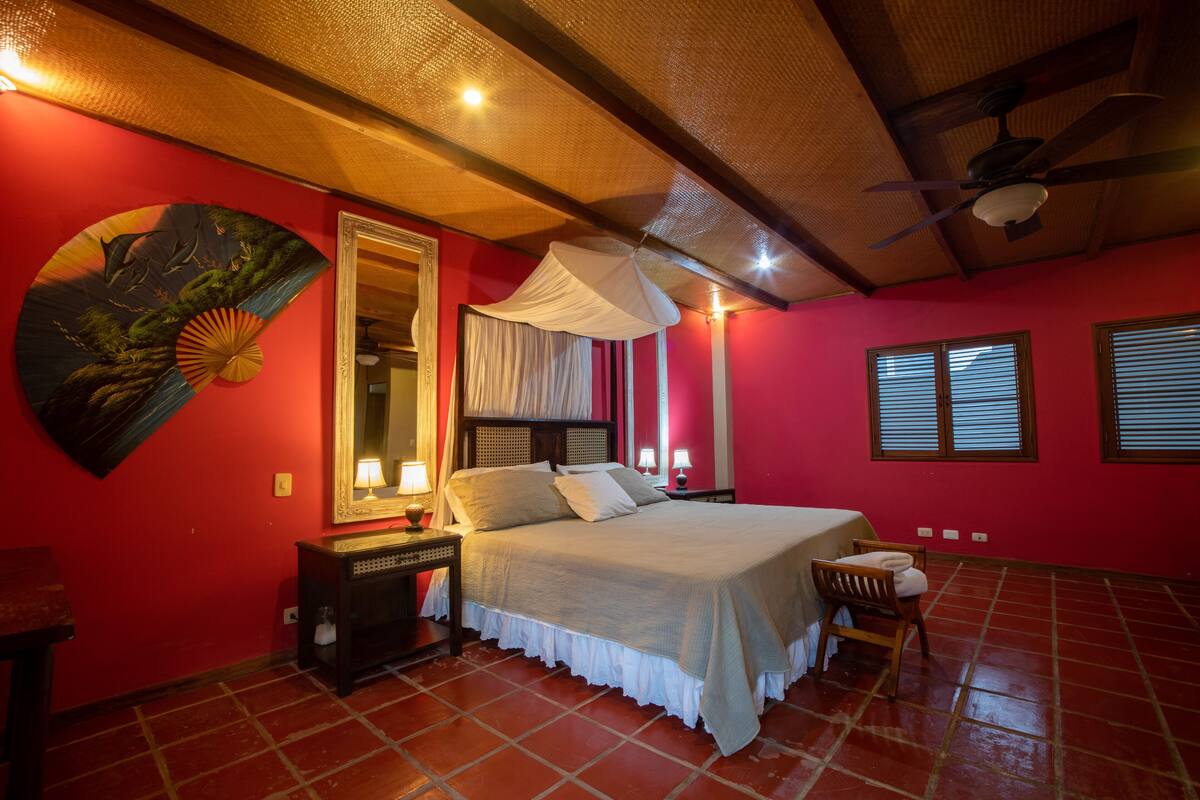 A bedroom with a bed, bedside tables, a mirror, a ceiling fan, and a red wall.