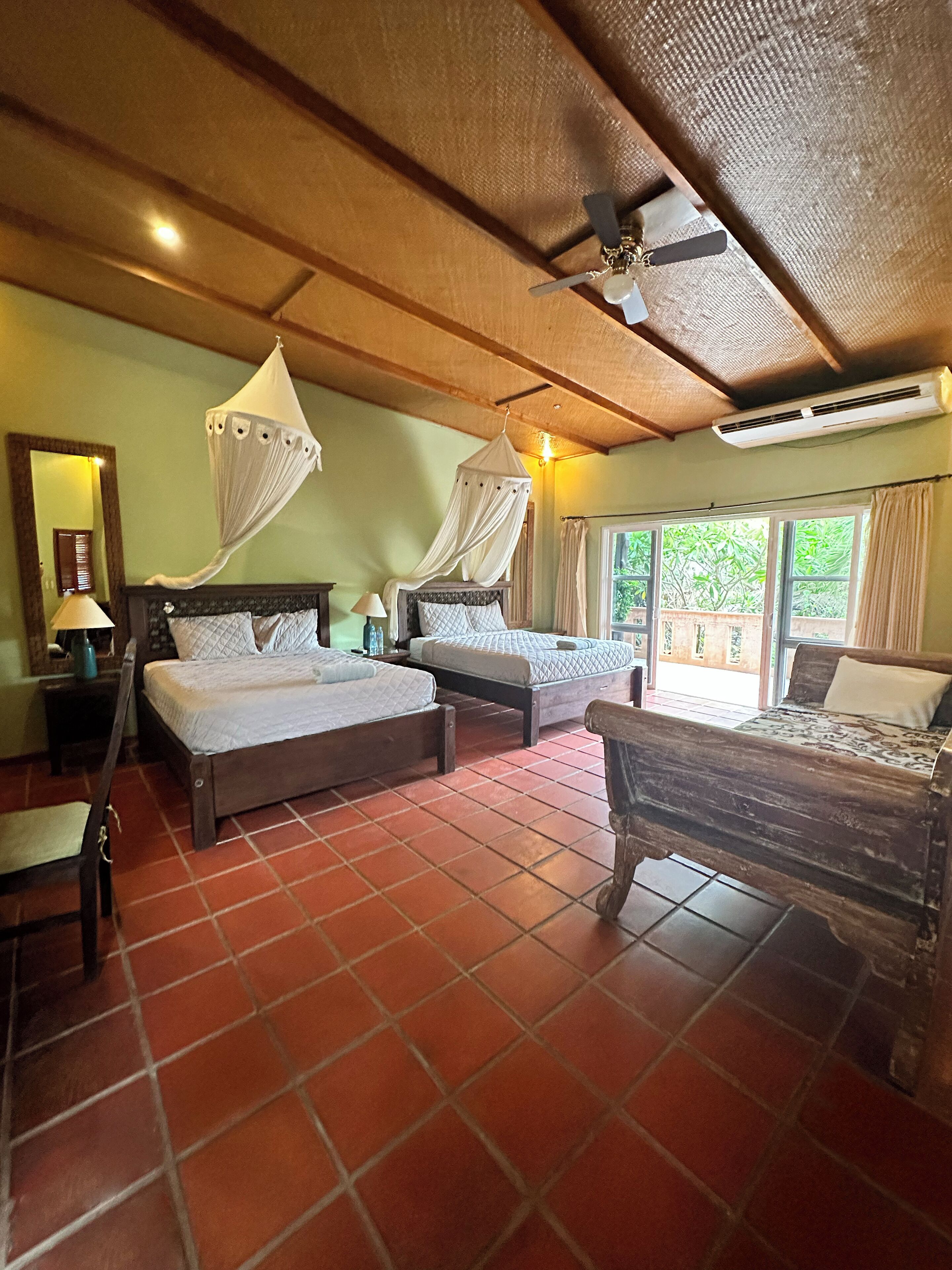 A room with two beds, a wooden ceiling, a ceiling fan, and a view of greenery outside.