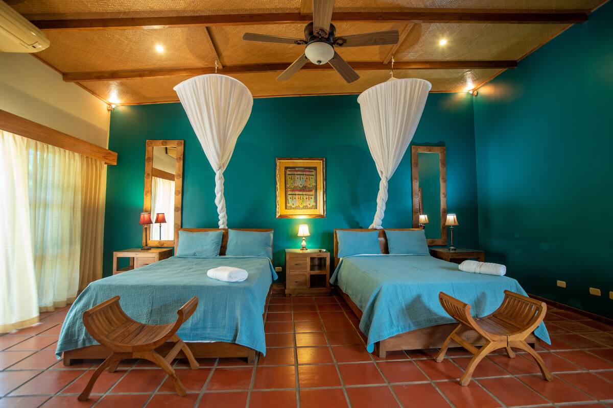 A room with two beds, wooden chairs, a ceiling fan, and a large mirror.